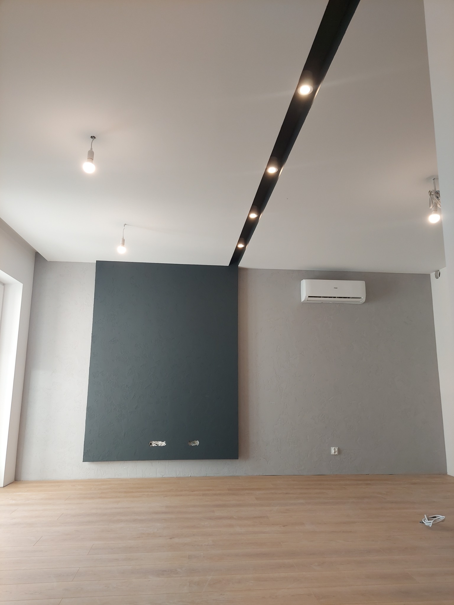 Pomalowane na szaro ściany w pokoju z drewnianą podłogą, z ciemnoszarą dekoracyjną wstawką na ścianie i klimatyzacją, widoczne także oświetlenie sufitowe.