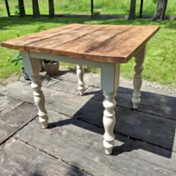 Kwadratowy stół z drewnianym blatem w odcieniach brązu i toczonymi, kremowymi nogami, ustawiony na drewnianym tarasie w słoneczny dzień.