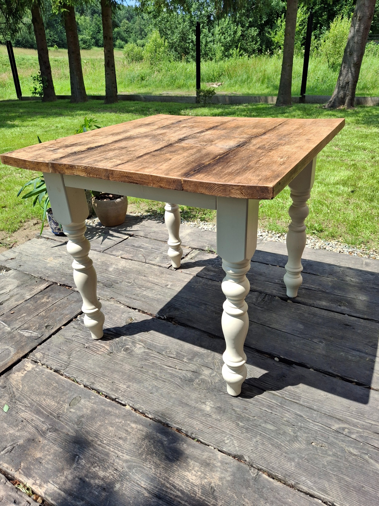 Kwadratowy stół z drewnianym blatem w odcieniach brązu i toczonymi, kremowymi nogami, ustawiony na drewnianym tarasie w słoneczny dzień.