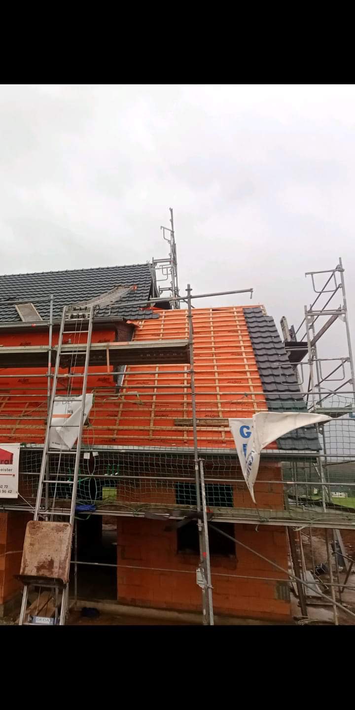 Montaż dachówki na nowym dachu, widoczne rusztowania, membrana dachowa i częściowo ułożone pokrycie w kolorze grafitowym, pochmurne niebo w tle.