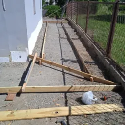 Przygotowanie podłoża pod wylewkę: drewniane szalunki ustawione wzdłuż ściany budynku i metalowej siatki ogrodzeniowej, widoczne narzędzia i materiały budowlane.