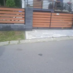 Nowoczesna brama przesuwna z poziomych paneli imitujących drewno, osadzona na szarych, betonowych słupkach, prowadząca do posesji z kostką brukową i zielonym trawnikiem.