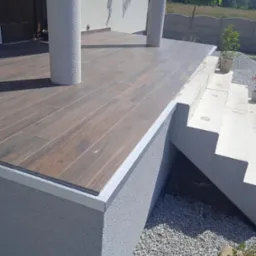 Wykończony taras z brązowych płytek imitujących drewno, obrzeże z metalową listwą, widoczne dwie okrągłe kolumny i schody z jasnych płyt betonowych.