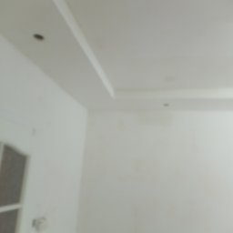 Ogólno budowlana - Niejasne ujęcie fragmentu pokoju z białym sufitem z listwami dekoracyjnymi i dwoma punktowymi lampami, oraz fragmentem białej ściany i drzwi z przeszkleniami.