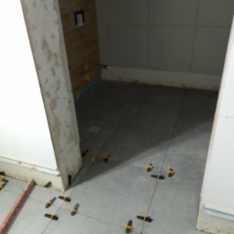 Ogólno budowlana - Układanie szarych płytek podłogowych w łazience z użyciem klipsów poziomujących, widoczne ślady zaprawy na płytkach i fugach, surowe wykończenie ściany w tle.