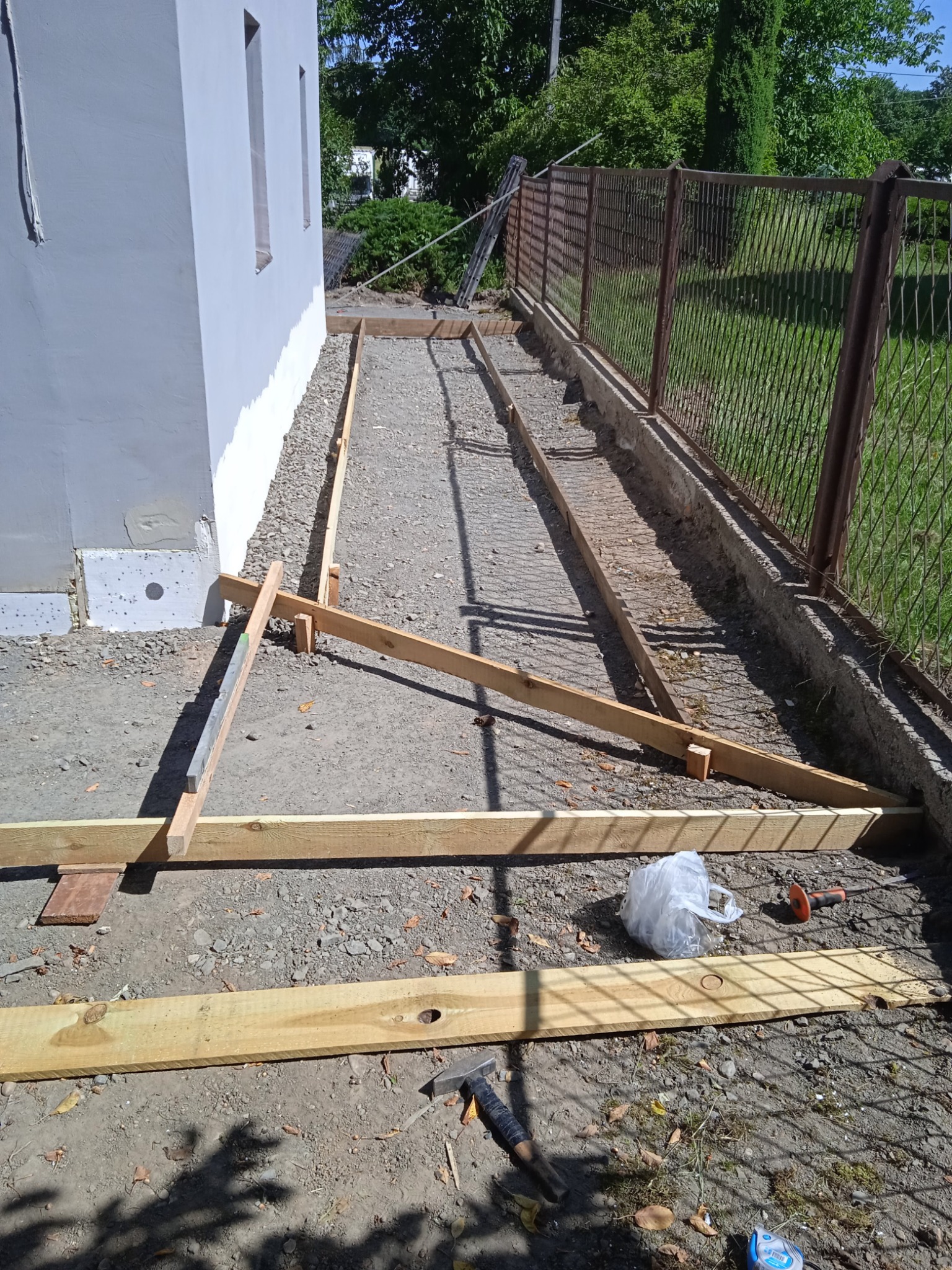 Przygotowanie podłoża pod wylewkę: drewniane szalunki ustawione wzdłuż ściany budynku i metalowej siatki ogrodzeniowej, widoczne narzędzia i materiały budowlane.