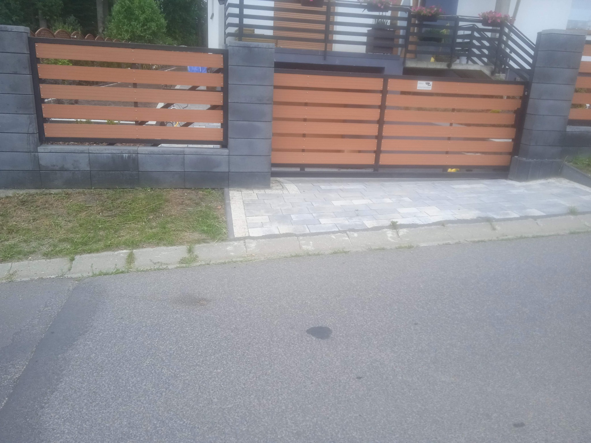 Nowoczesna brama przesuwna z poziomych paneli imitujących drewno, osadzona na szarych, betonowych słupkach, prowadząca do posesji z kostką brukową i zielonym trawnikiem.