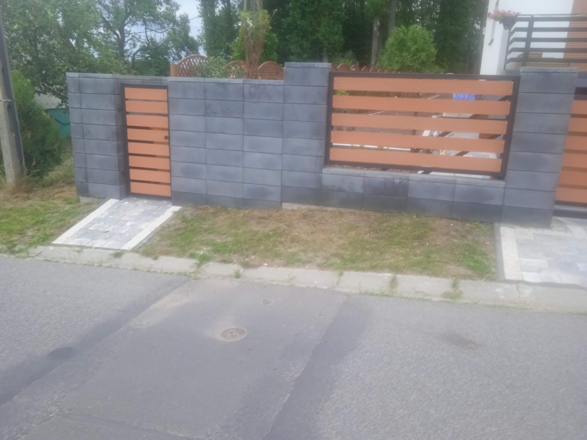 Nowoczesne ogrodzenie posesji z szarych betonowych bloczków i poziomych paneli z tworzywa imitującego drewno, z furtką i fragmentem chodnika z kostki brukowej.