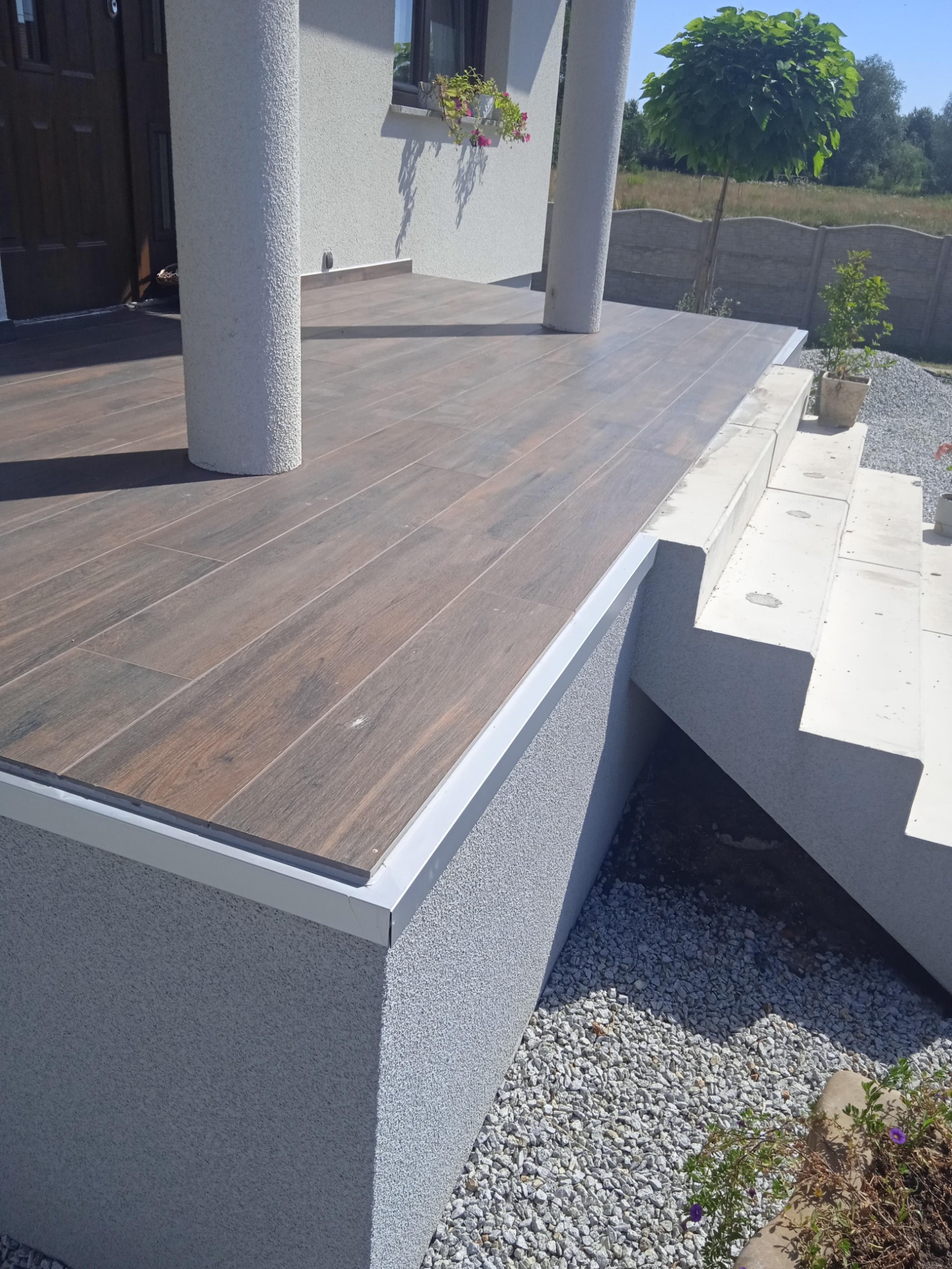 Zadaszony taras z brązowymi płytkami imitującymi drewno, obudowany szarym tynkiem strukturalnym i wykończony srebrną listwą, z kolumnami i schodami prowadzącymi do ogrodu.