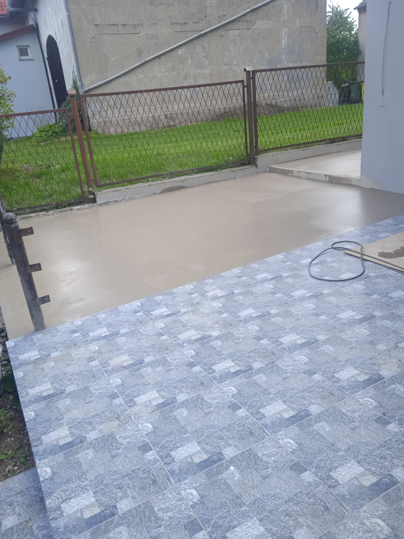 Świeżo wylana wylewka betonowa na tarasie sąsiaduje z ułożoną szarą, wzorzystą terakotą. W tle widoczne ogrodzenie i budynek.