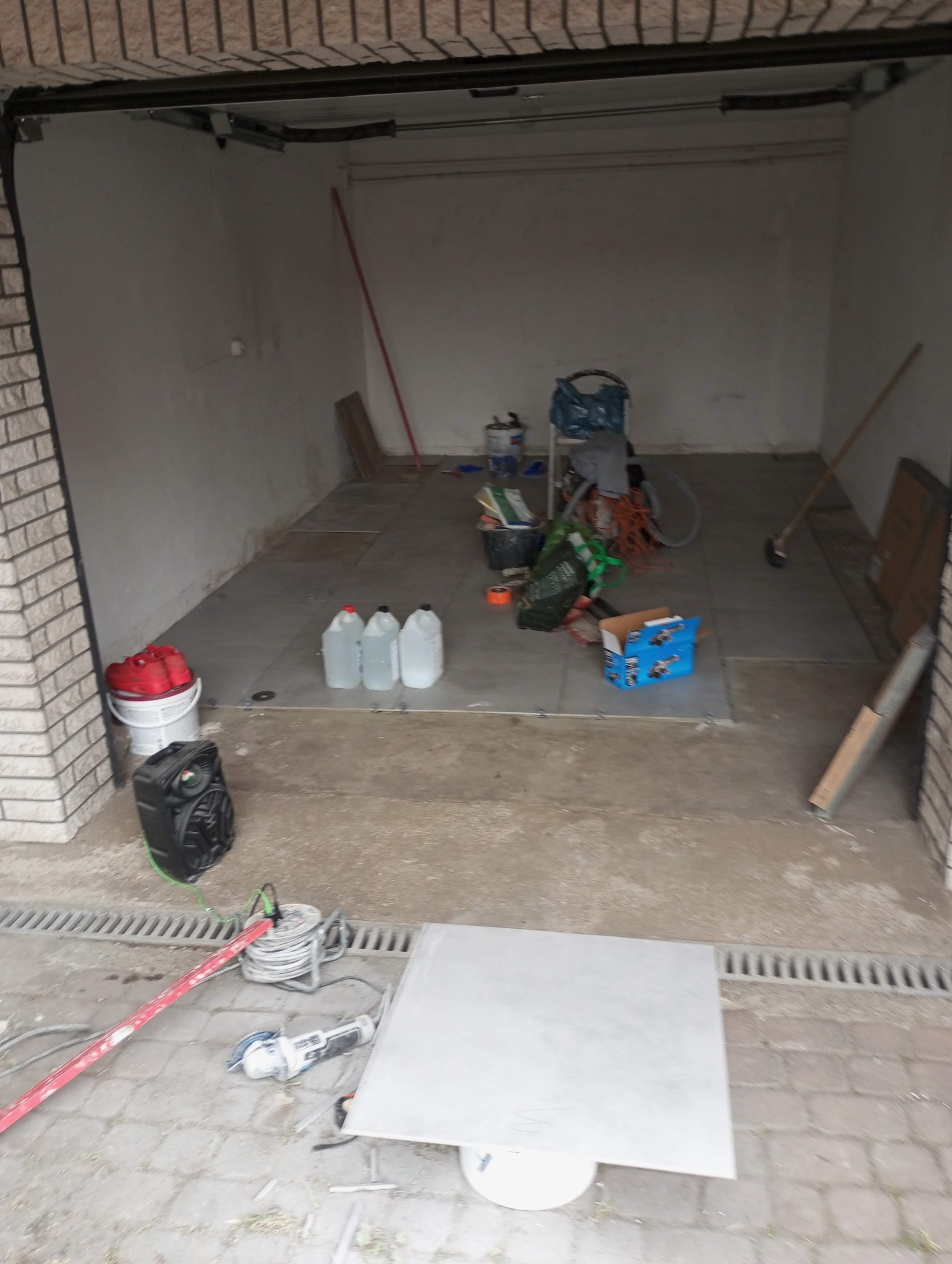 Garaż w trakcie układania szarych płytek podłogowych, widoczne narzędzia glazurnicze, poziomica, zapas płytek, pojemniki z płynami, oraz krzyżyki dystansowe.
