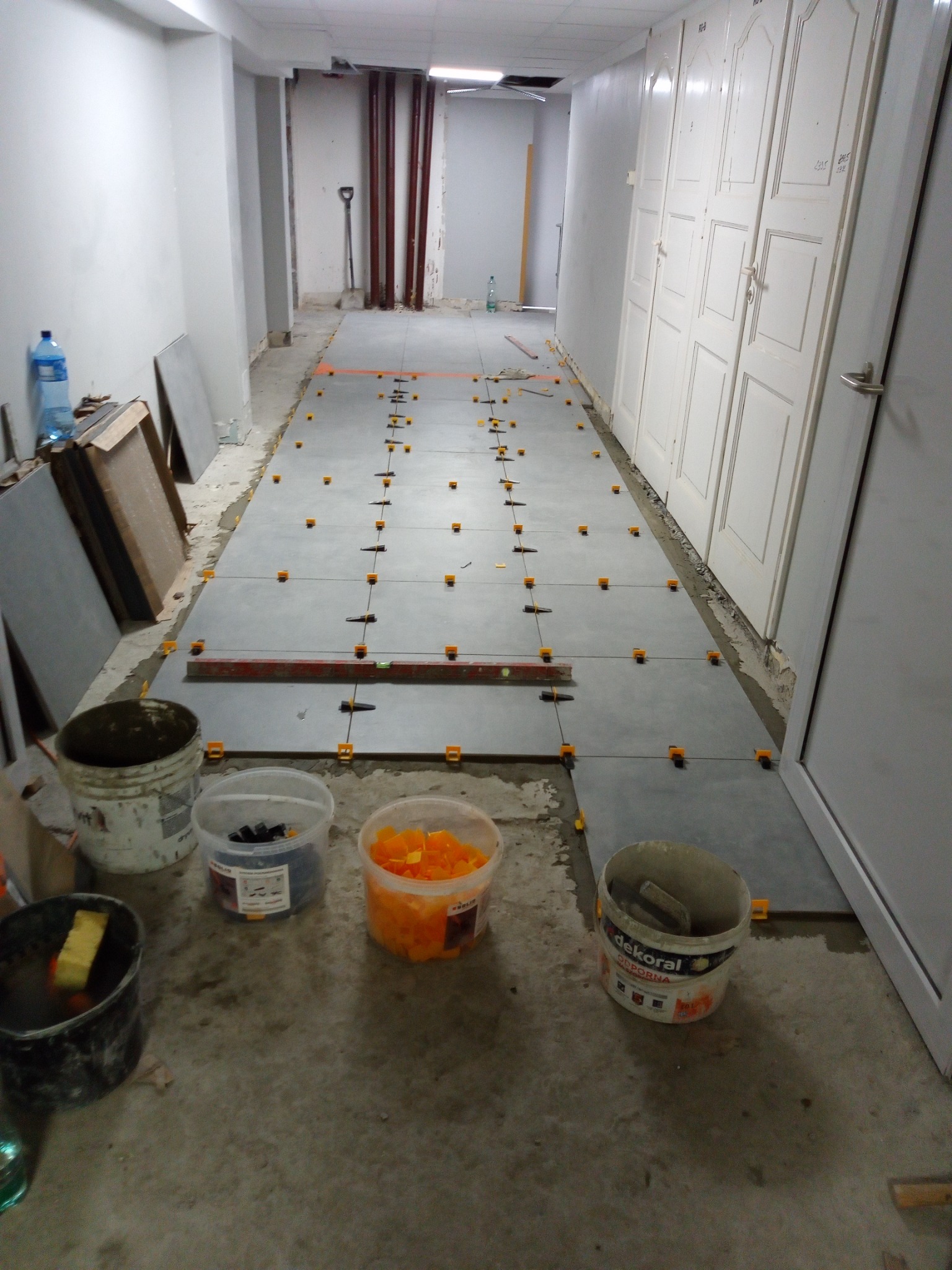 Korytarz w trakcie układania szarych płytek podłogowych z użyciem poziomicy i pomarańczowych krzyżyków dystansowych, widoczne pojemniki z akcesoriami i narzędzia.