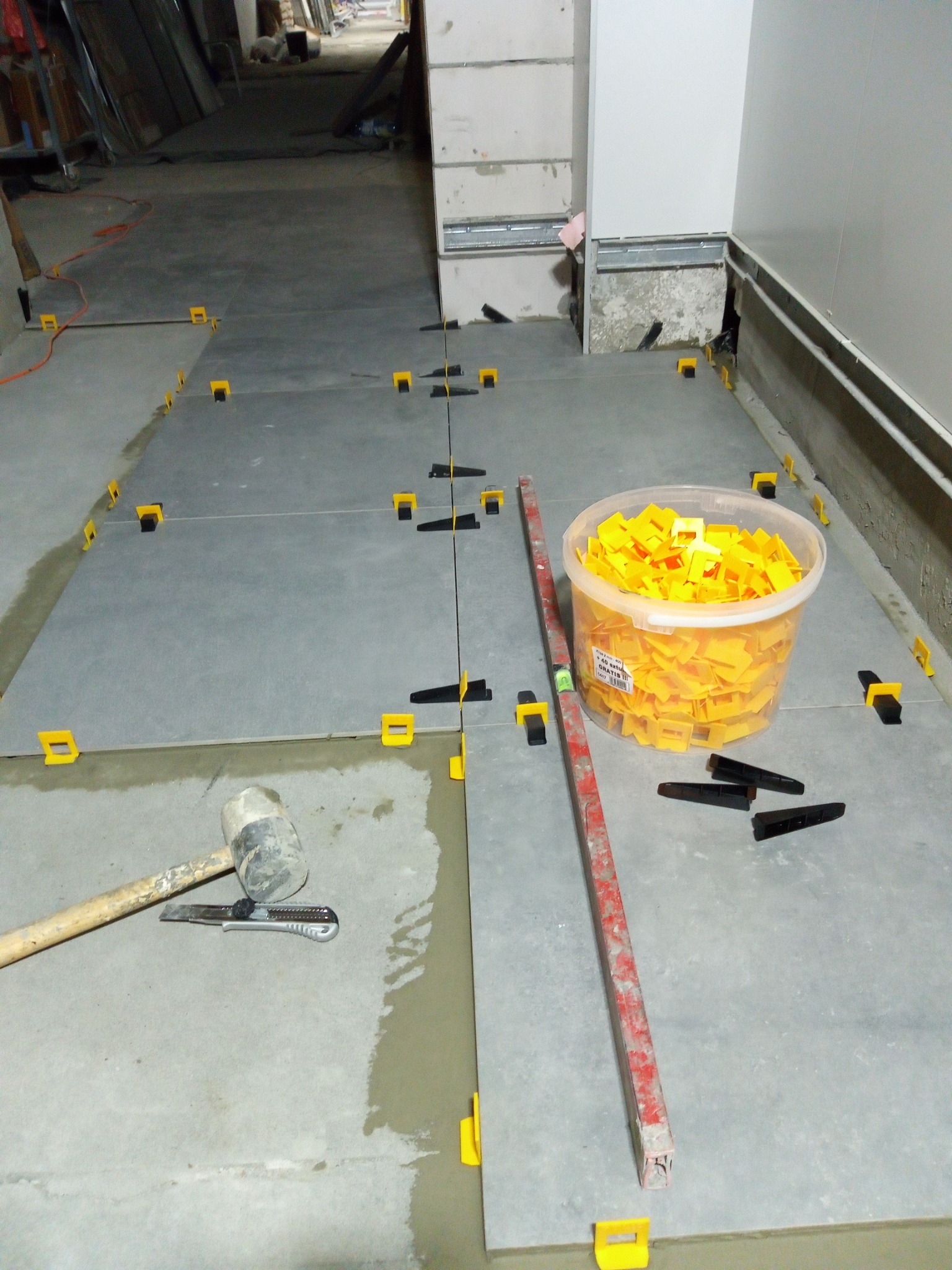 Szare płytki podłogowe w trakcie układania, z użyciem poziomicy, gumowego młotka, nożyka i systemu poziomowania płytek (klipsy i kliny).