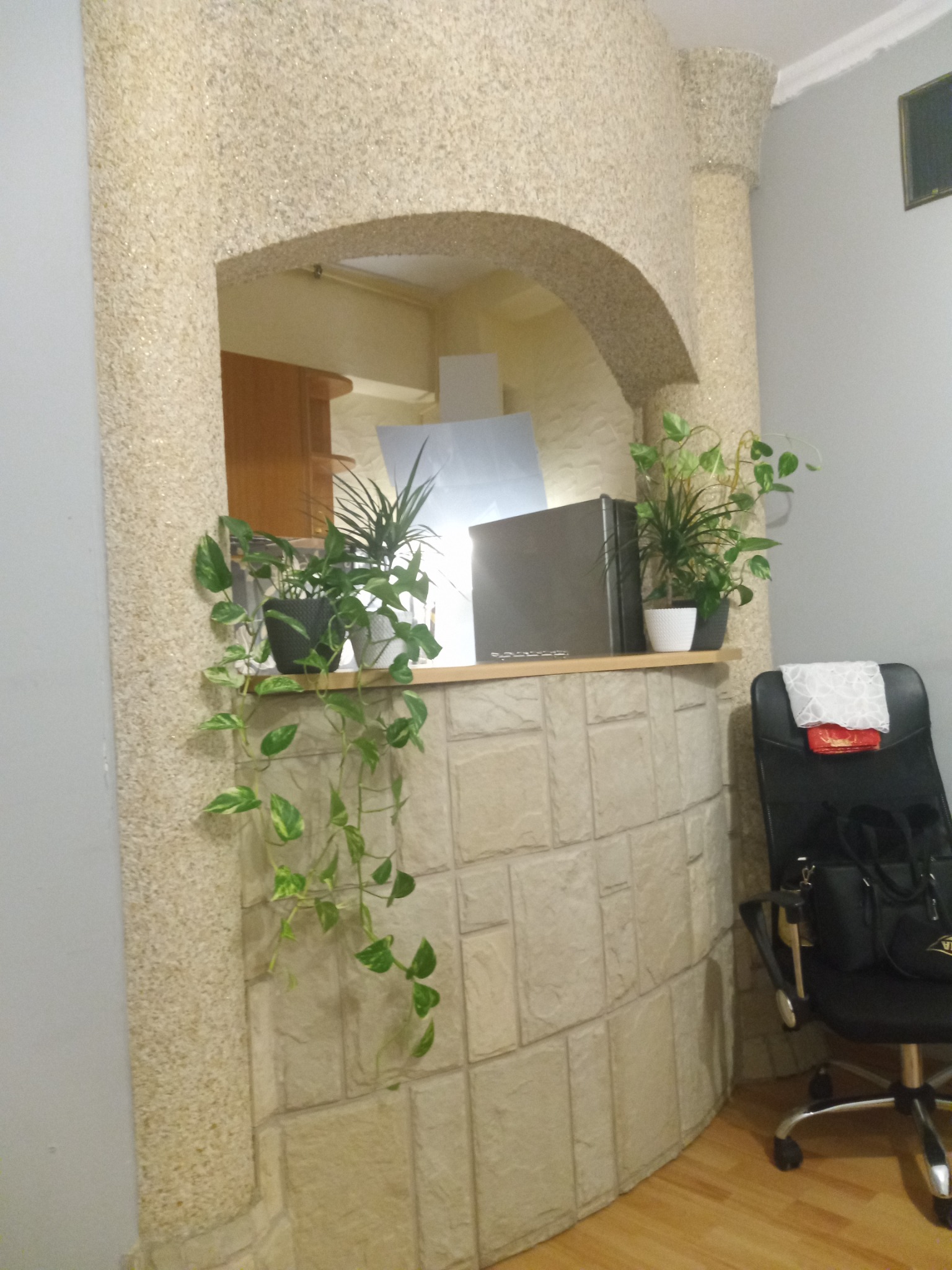 Aranżacja wnętrza z łukowym przejściem wykończonym dekoracyjnym tynkiem strukturalnym, na półce rośliny doniczkowe i sprzęt AGD, obok krzesło biurowe.