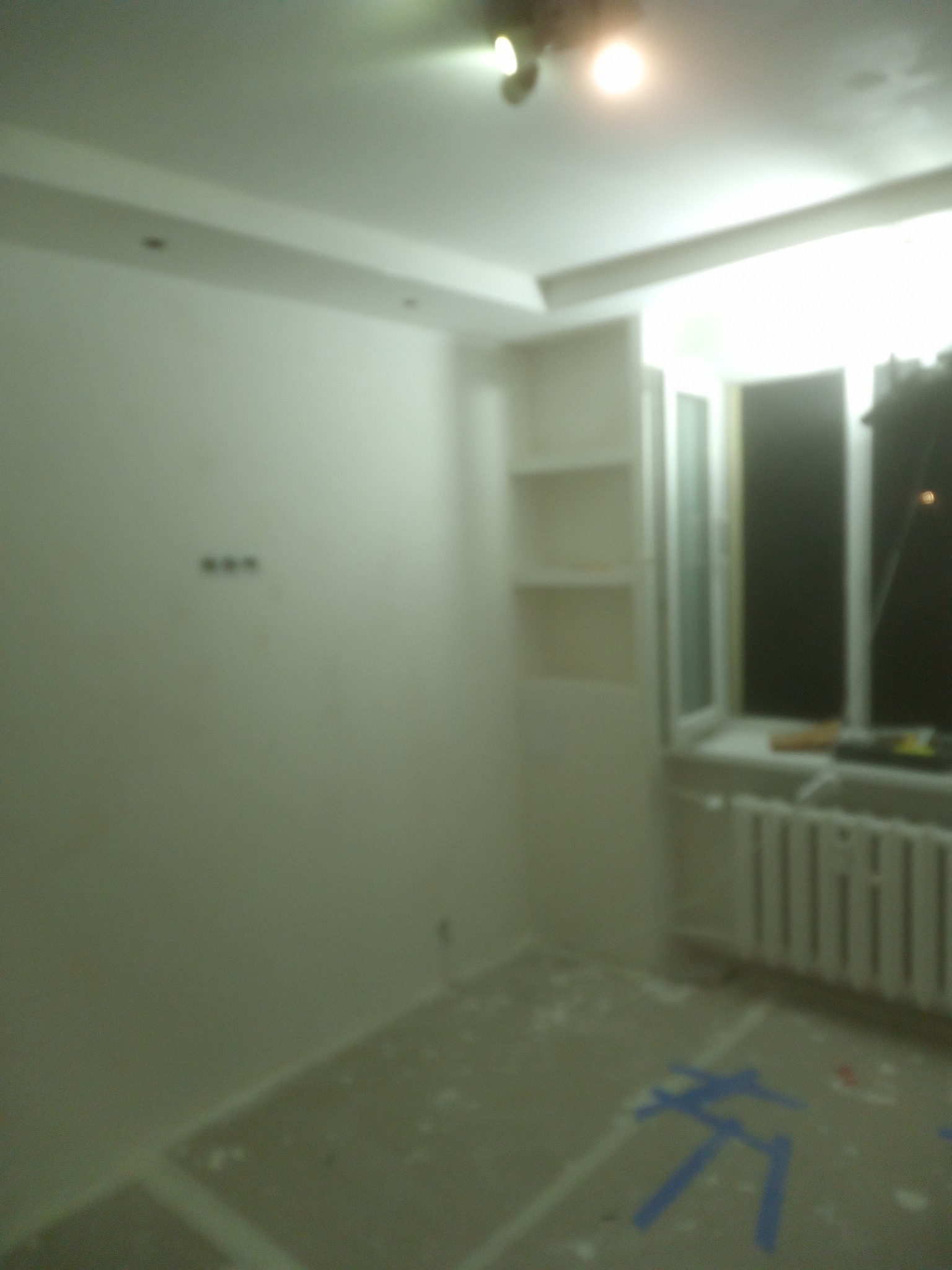 Pokój w trakcie remontu: świeżo pomalowane ściany, surowy beton na podłodze z niebieską taśmą malarską, wnęka z półkami do zabudowy obok okna z grzejnikiem.