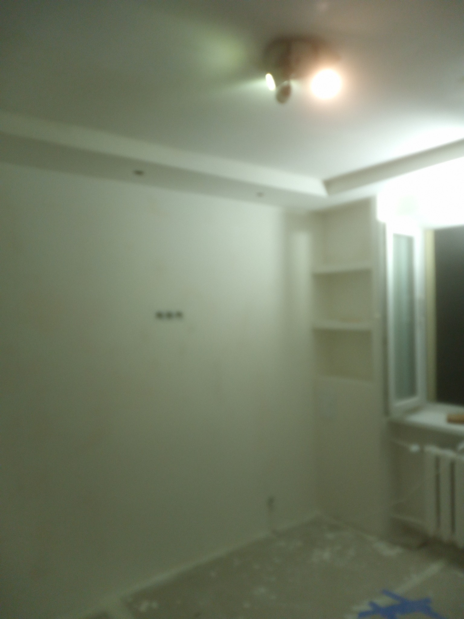 Pokój po malowaniu ścian na biało, widoczne ślady farby na podłodze, grzejnik pod oknem i wnęka z półkami w ścianie.