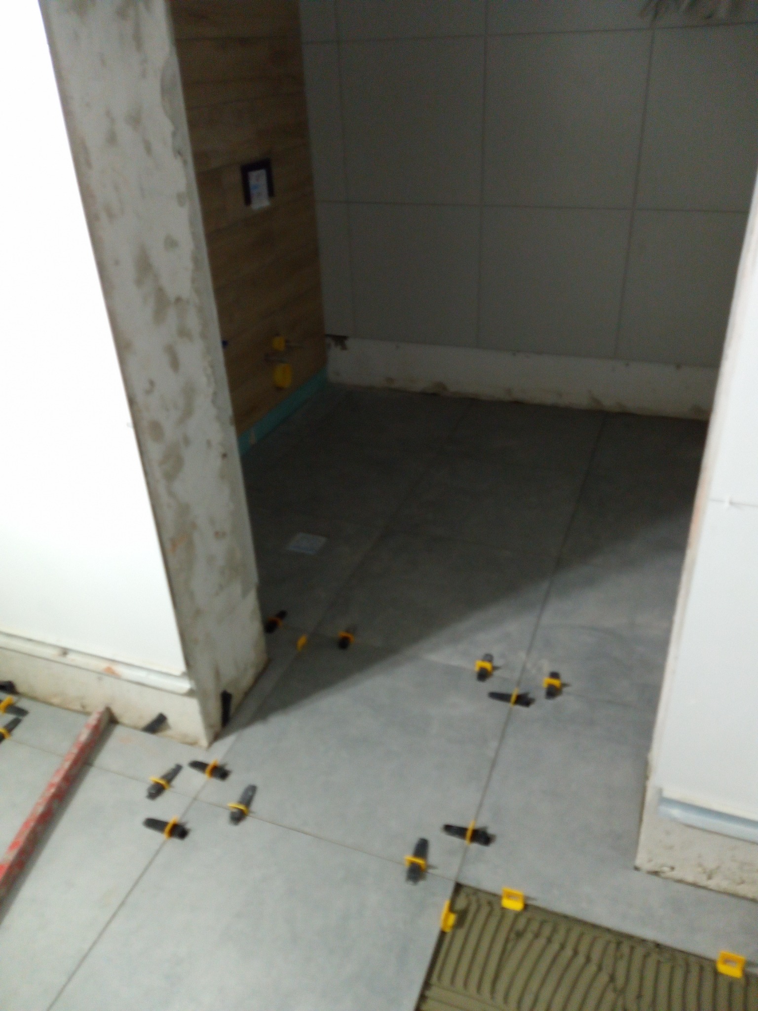 Układanie szarych płytek podłogowych w łazience z użyciem klipsów poziomujących, widoczne ślady zaprawy na płytkach i fugach, surowe wykończenie ściany w tle.