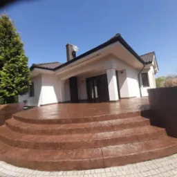 Elewacja domu jednorodzinnego z brązowym, stemplowanym betonem na schodach wejściowych i tarasie, widok z dołu na tle błękitnego nieba.