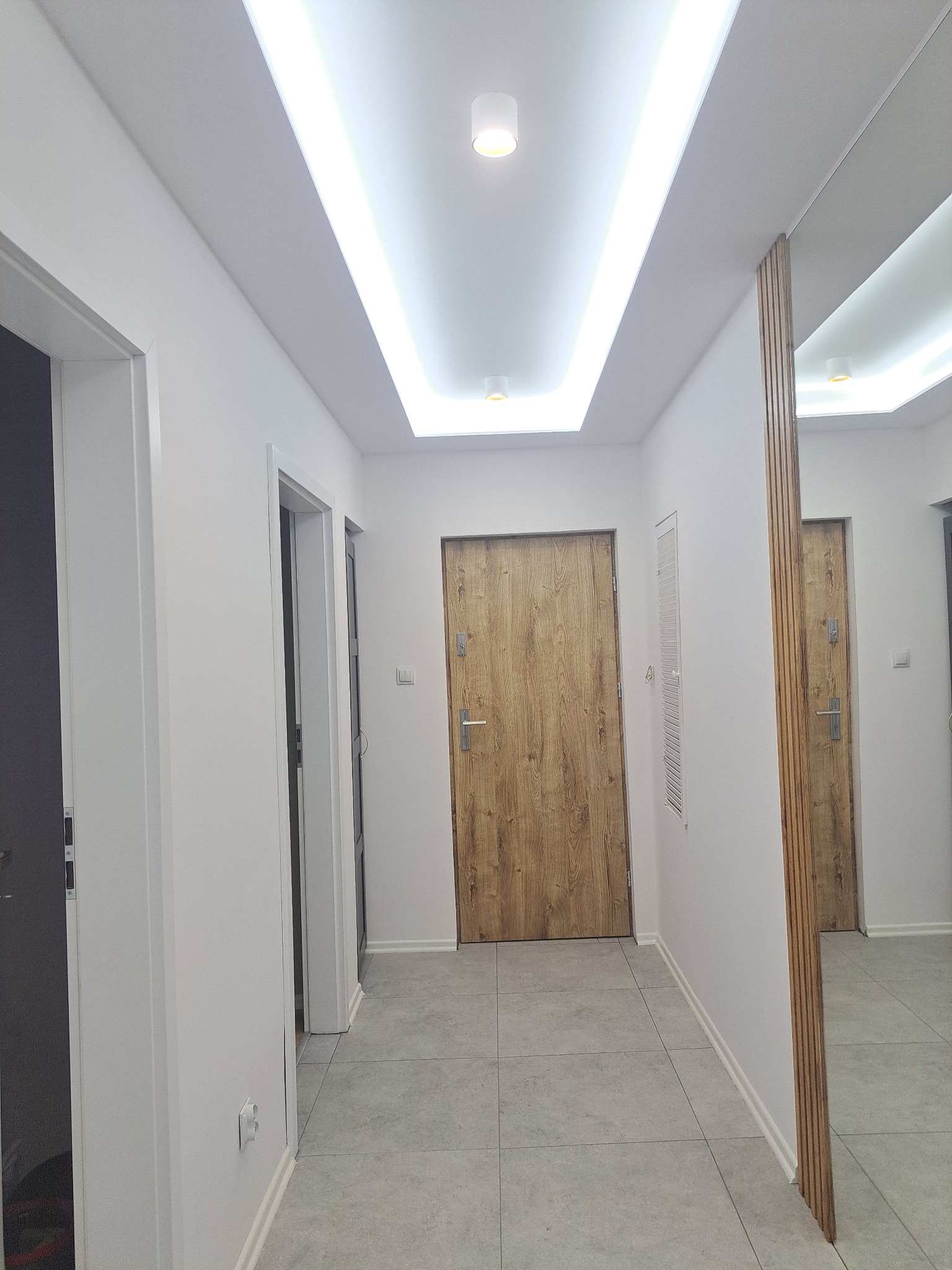 Nowoczesny korytarz z drewnianymi drzwiami, oświetleniem LED na suficie i dużym lustrem z drewnianymi listwami. Szare płytki podłogowe i białe ściany.