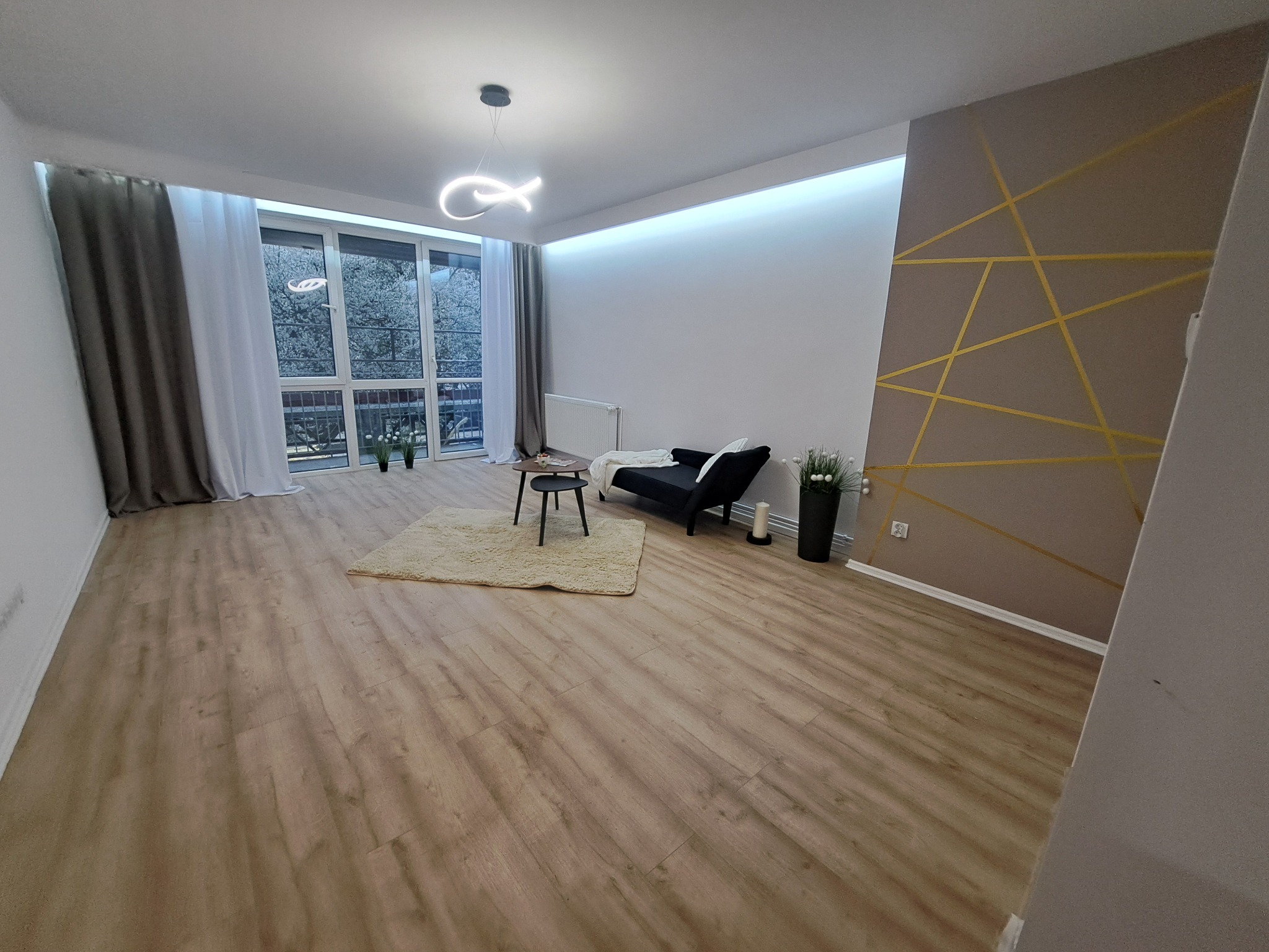 Jasne wnętrze po wykończeniu: panele podłogowe, ściana z geometrycznym wzorem w odcieniach beżu i złota, okno z widokiem na zieleń, nowoczesne oświetlenie.