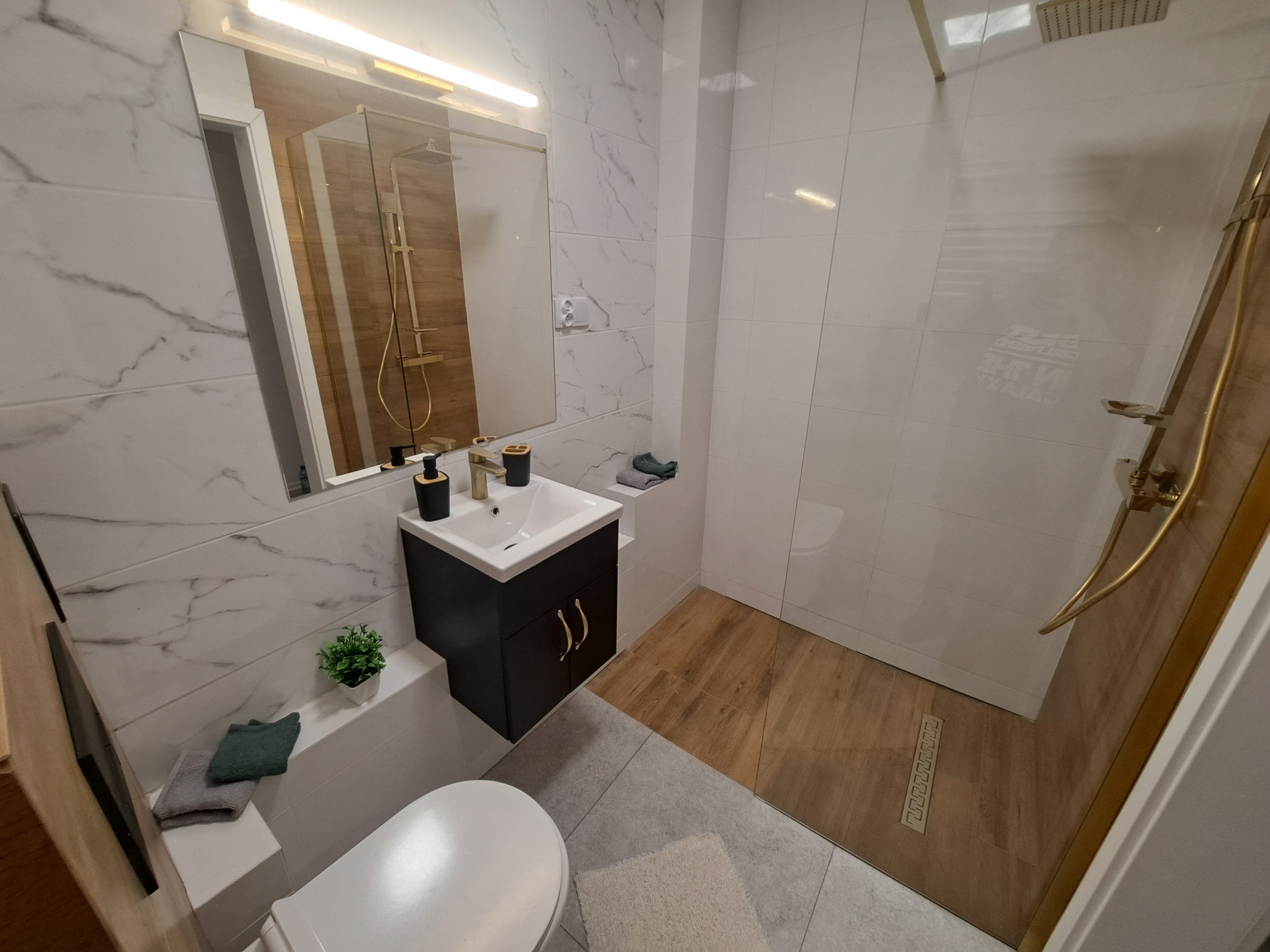 Nowoczesna łazienka z marmurowymi płytkami, kabiną prysznicową typu walk-in i czarną szafką pod umywalką z złotymi uchwytami. Widoczny fragment toalety i dekoracje.