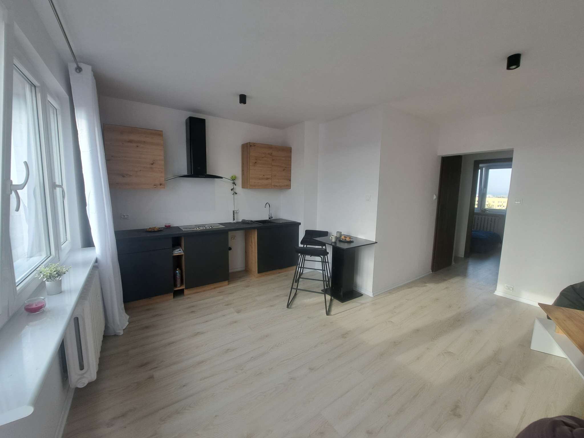 Nowoczesna kuchnia z czarnymi szafkami i drewnianymi akcentami, jasne panele podłogowe, stół barowy z krzesłami. Widok na okno z białymi zasłonami.