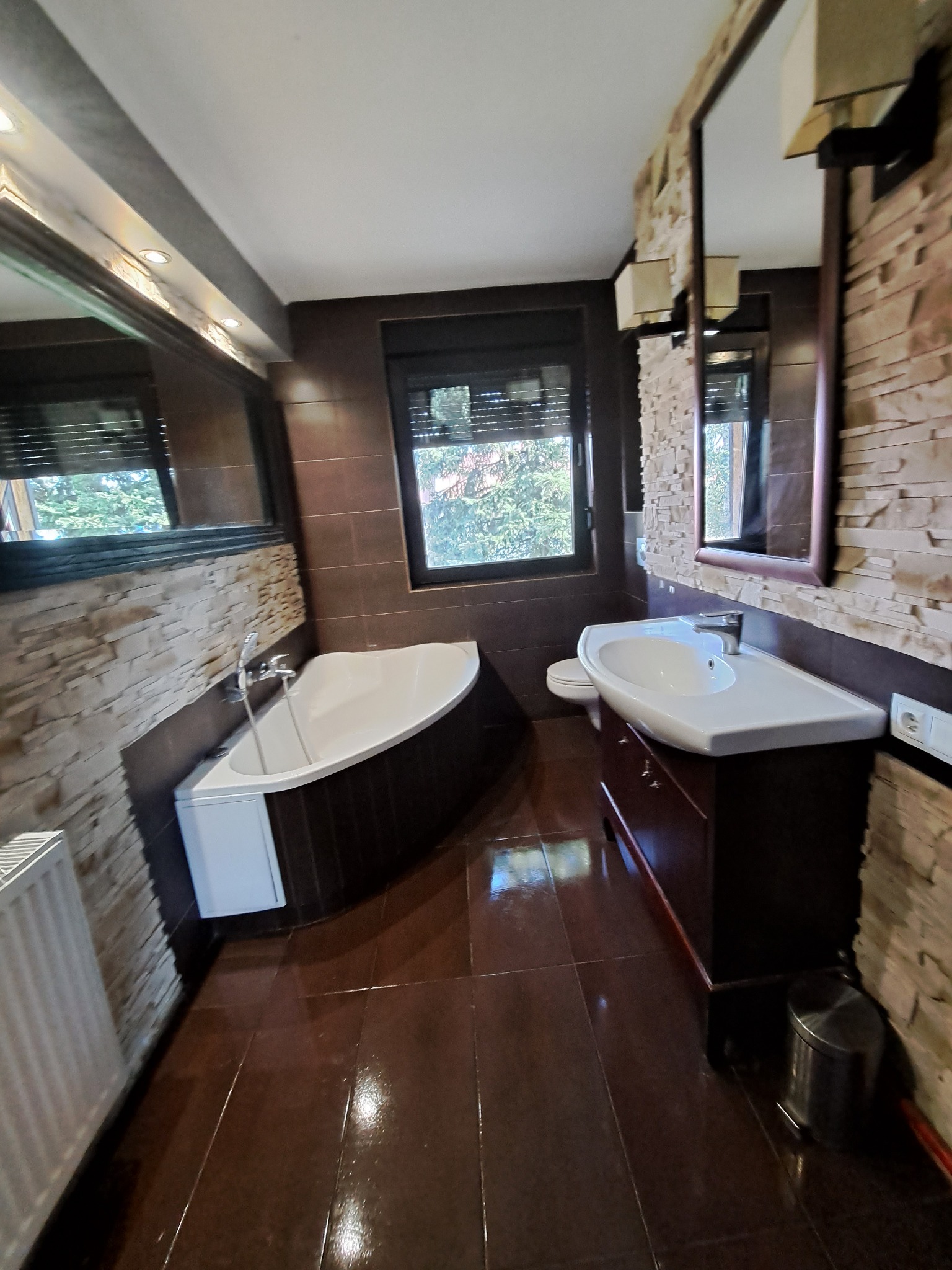 Nowoczesna łazienka z wanną narożną, umywalką na szafce z ciemnego drewna i ścianami wykończonymi kamieniem dekoracyjnym. Okno z żaluzjami w tle.