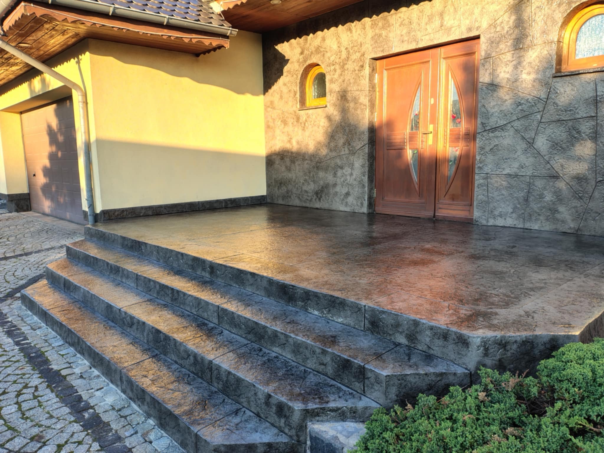 Wejście do domu z czterostopniowymi schodami wyłożonymi szarą kostką, z widoczną elewacją w odcieniach beżu i szarości oraz drewnianymi drzwiami wejściowymi.