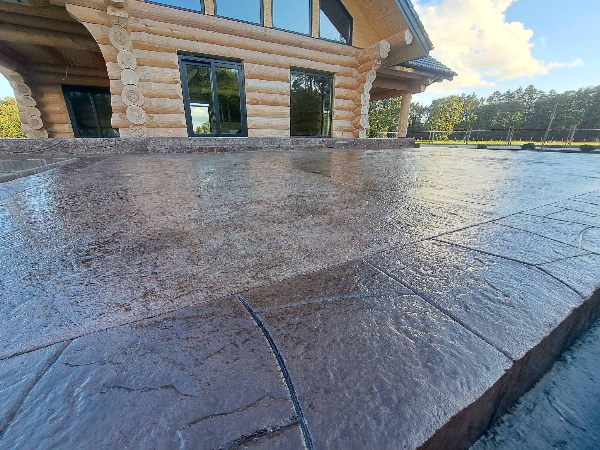 Imponujący taras z betonu stemplowanego przy drewnianym domu z bali, z widoczną fakturą kamienia i eleganckim wykończeniem krawędzi. Całość w stonowanej kolorystyce.