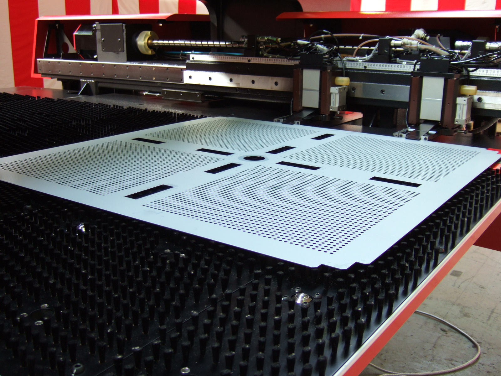 Maszyna CNC perforuje arkusz blachy z otworami i wycięciami, leżący na czarnym podłożu z pinami.