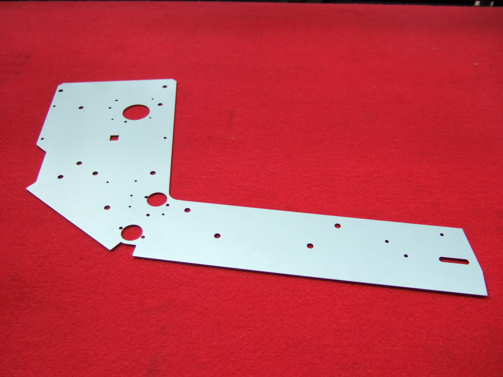 Metalowy element obudowy urządzenia wentylacyjnego lub klimatyzacyjnego z otworami o różnych średnicach, leżący na czerwonym tle.