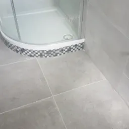 Szare, kwadratowe płytki podłogowe i ścienne w łazience z kabiną prysznicową z mozaikowym wykończeniem, widoczne dwa chromowane zawory na ścianie.