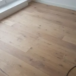 Jasna, drewniana podłoga ułożona w nowym, pustym pokoju z białymi ścianami, listwami przypodłogowymi i oknem.