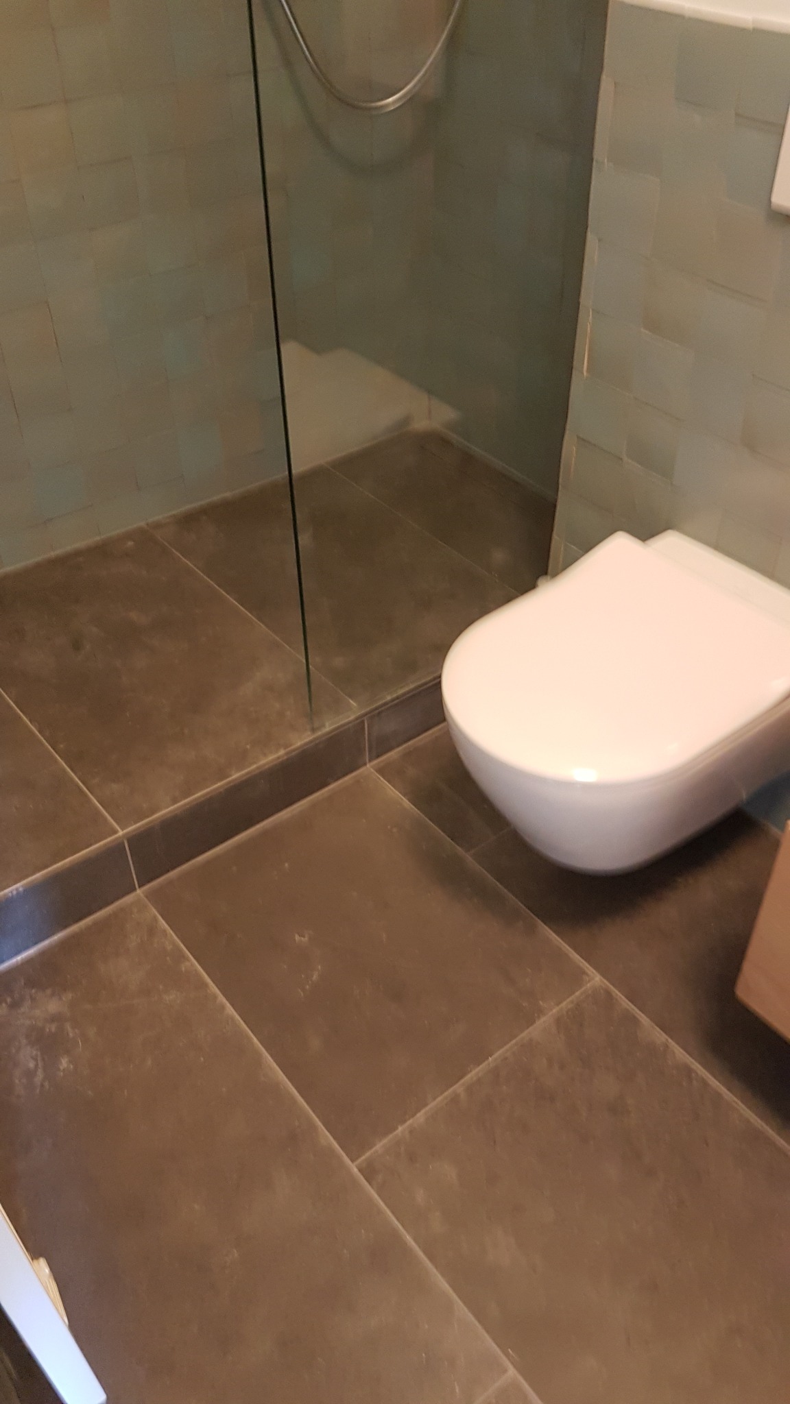 Nowoczesna łazienka z brązowymi płytkami podłogowymi, szklaną kabiną prysznicową i podwieszaną toaletą.