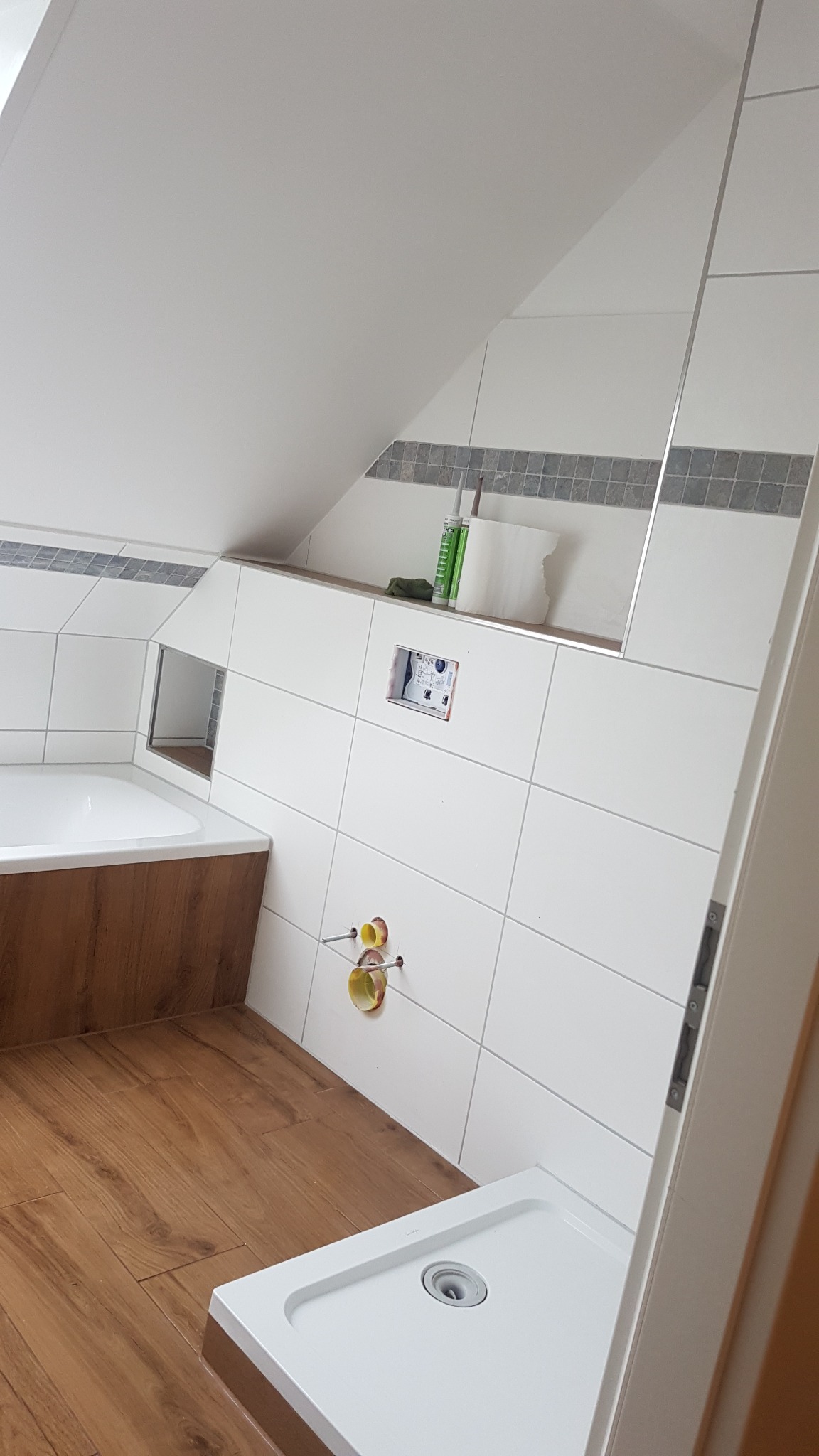 Poddasze łazienkowe z białą glazurą, drewnopodobną podłogą i brodzikiem; widoczne przygotowane instalacje wodne i półka z akcesoriami.