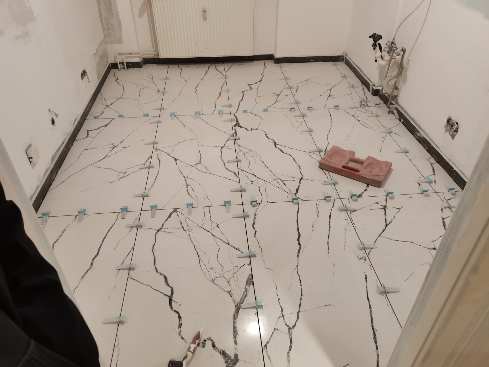 Układanie białych płytek podłogowych z efektem marmuru, używając niebieskich krzyżyków dystansowych, widoczny czerwony gumowy młotek do dobijania płytek i fragment instalacji wodnej przy ścianie.