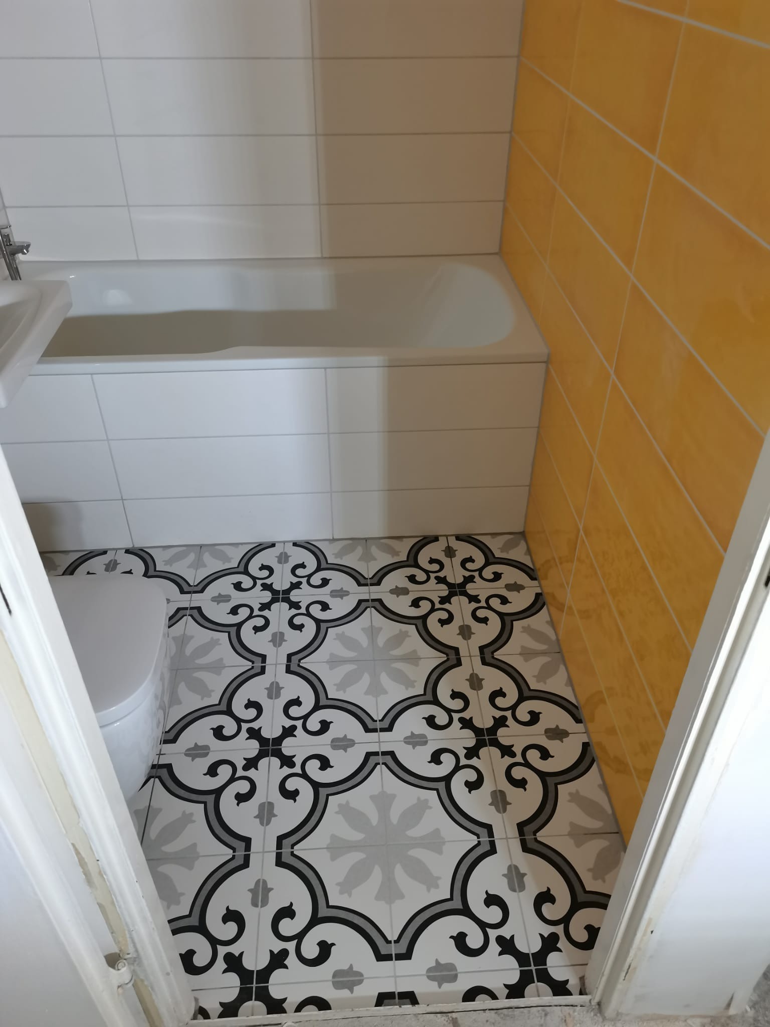 Biała wanna i toaleta w łazience z żółtymi płytkami na ścianie i wzorzystą podłogą z płytek w odcieniach szarości i czerni, widok zza uchylonych drzwi.
