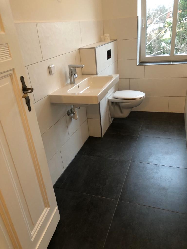 Nowoczesna łazienka z białymi płytkami ściennymi, wiszącą umywalką z chromowaną baterią, toaletą oraz ciemną, matową podłogą z płytek. Widoczny fragment białych, drewnianych drzwi.