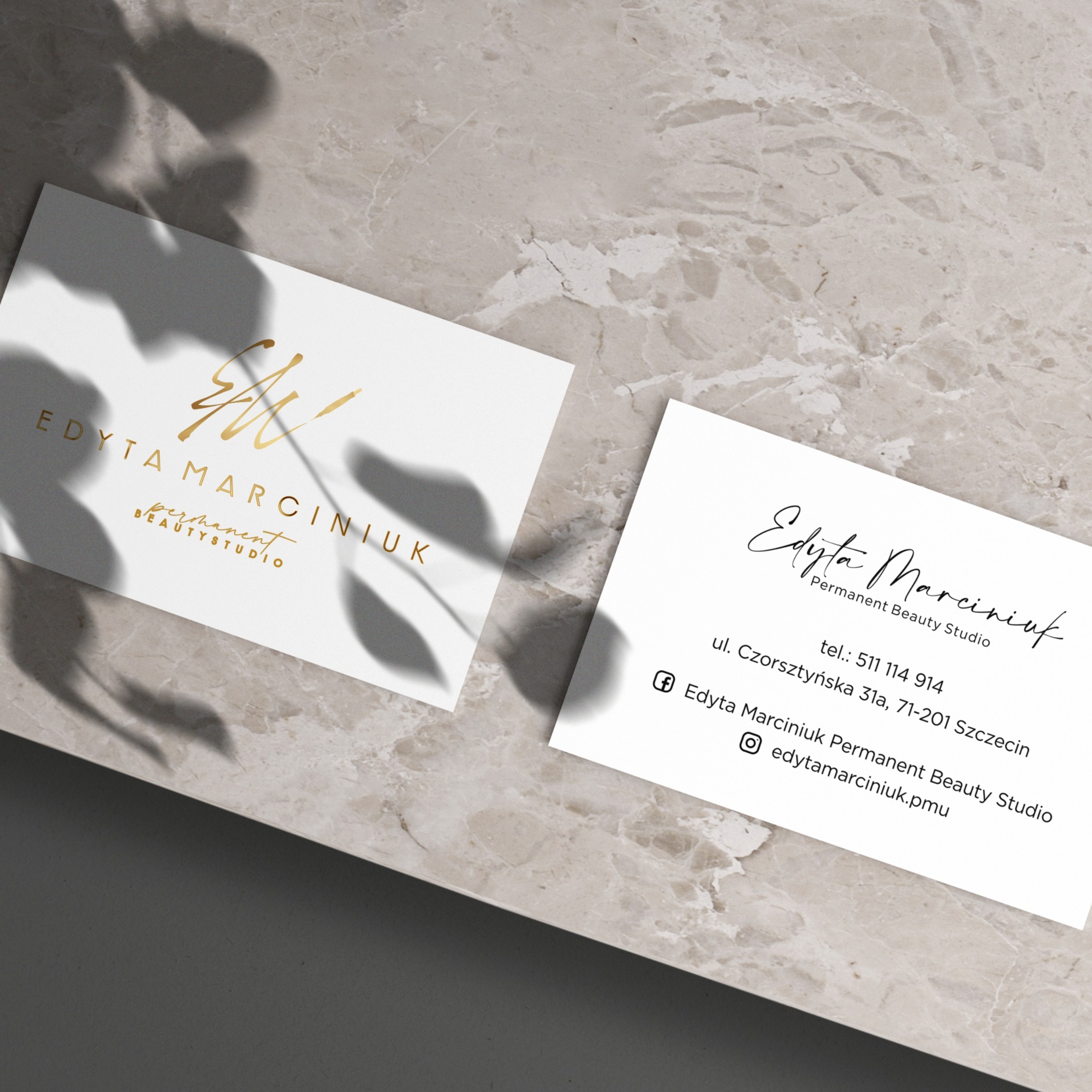 Dwie wizytówki na marmurowym tle, jedna z logo w kolorze złotym, druga z czarnym tekstem zawierającym dane kontaktowe, cień liści rzucający się na wizytówki.