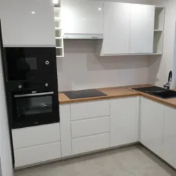 Nowoczesna, biała kuchnia z drewnianym blatem, zabudowanym piekarnikiem i mikrofalówką marki Amica, z widoczną płytą indukcyjną i czarnym zlewozmywakiem.