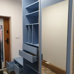 Niebieska szafa wnękowa z otwartymi szufladami i lustrem, widoczny wzorzysty parkiet i częściowo zamontowana ściana.