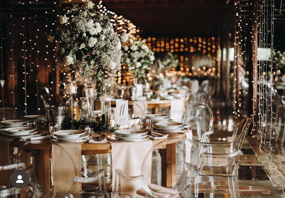 Elegancko nakryty stół z numerem 11, kwiatowymi dekoracjami, świecami i przezroczystymi krzesłami, oświetlony girlandami świetlnymi w tle.