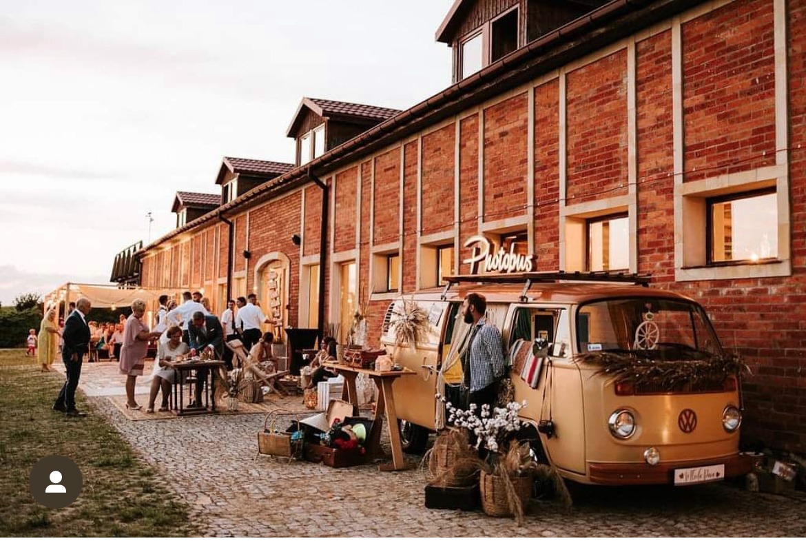 Beżowy VW Transporter przerobiony na fotobudkę 'Photobus' na tle ceglanego budynku podczas imprezy plenerowej, goście w tle.