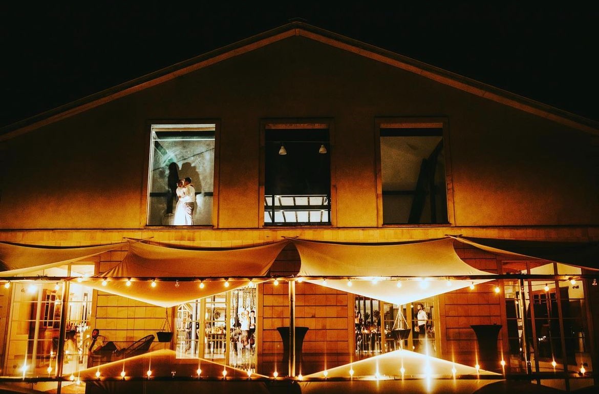 Elewacja budynku z oknami, w jednym z nich para w objęciach, na dole taras z oświetleniem i gośćmi, nocna sceneria.