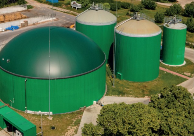 Widok z góry na biogazownię z czterema zielonymi zbiornikami o różnej wielkości, z których jeden ma kopułę, otoczoną zielenią i elementami infrastruktury.