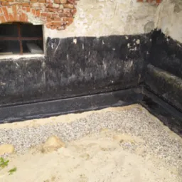Fundament zabezpieczony czarną izolacją przeciwwodną, drenaż wzdłuż ściany, piasek i żwir na podłożu, okno z ceglanym obramowaniem w tle.