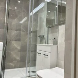 Nowoczesna łazienka z kabiną prysznicową ze szklanymi drzwiami, umywalką z szafką i toaletą, wykończona szarymi płytkami imitującymi marmur, oświetlona listwą LED nad lustrem.