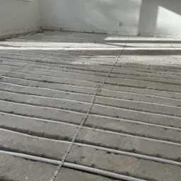 Białe rurki grzewcze ułożone równolegle na szarym podłożu w trakcie instalacji, z widoczną listwą przypodłogową i fragmentem okna w tle.