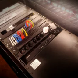 Widok z góry na szafę rack z zamontowanymi urządzeniami, w tym listwą zasilającą, zasilaczami i okablowaniem w kolorowych peszlach.