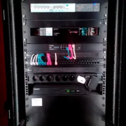 Szafa rack z zainstalowanymi urządzeniami sieciowymi, zasilaczami i centralą alarmową Hikvision, kable uporządkowane kolorowymi opaskami.